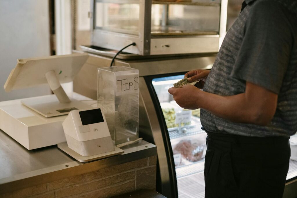 Unrecognizable male in casual clothes standing with money near cash register in grocery store while making purchase near glass showcase